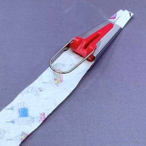 May include: A red and silver fabric pressing tool with a metal loop handle. The tool is pressing a white fabric with a colorful pattern.