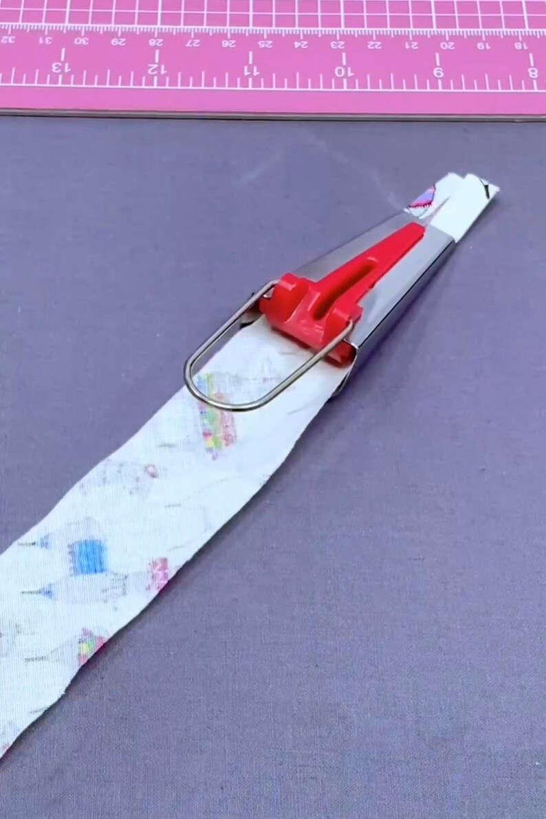 May include: A red and silver fabric pressing tool with a metal loop handle. The tool is pressing a white fabric with a colorful pattern.