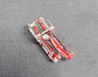 Puede incluir: Un pie de máquina de coser de plástico transparente con marcas rojas. El pie tiene un tornillo de metal para sujetarlo a la máquina de coser.