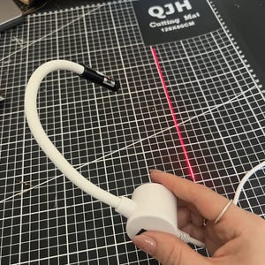 May include: A white laser light with a flexible neck and a black and silver tip. The laser emits a bright red line, and the device is held over a black cutting mat with white grid lines. The mat has the text "QJH Cutting Mat 120x60CM".