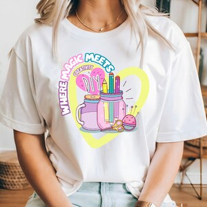 May include: White t-shirt with a graphic design of two jars filled with colorful pencils and scissors. The text "Where Magic Meets Creativity DIY" is written in a circle around the jars.