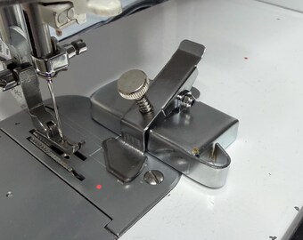 May include: Close-up of a sewing machine needle and presser foot. The machine is silver and white, with a silver metal attachment. The attachment has a screw and a hinged arm. The needle is positioned to sew.