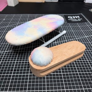 May include: A small ironing board with a pastel tie-dye cover and a wooden seam roll. The ironing board has a white metal frame. The seam roll has a light wood base and a pastel tie-dye cushion.