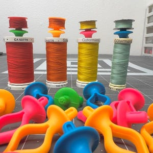 Peut inclure: Une collection de bobines de fil de couleurs vives avec des bobines et des guides-fils. Les bobines sont orange, jaune et bleue. Les bobines sont blanches et les guides-fils sont verts. Il y a aussi plusieurs porte-fils en plastique de couleurs vives.