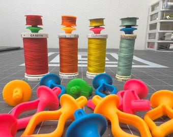 May include: A collection of colorful thread spools with bobbins and thread guides. The spools are orange, yellow, and blue. The bobbins are white and the thread guides are green. There are also several colorful plastic thread holders.