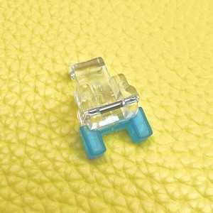 May include: A clear plastic sewing machine foot with two blue plastic pieces on the sides. The foot is designed for sewing on a sewing machine.