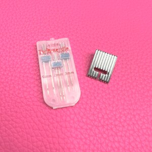 May include: A clear plastic case containing twin needles for sewing. The case has the text "Twin needle" and "Size 2mm 3mm" printed on it. The needles are silver and come in three different sizes. A silver metal sewing machine foot is also pictured.