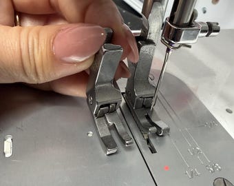 Könnte beinhalten: Nahaufnahme einer Nähmaschine mit zwei Metall-Nähfüßen. Die Nadel ist sichtbar, und das Maschinenbett hat Messmarkierungen. Das Bild zeigt eine Nähmaschine in Gebrauch.