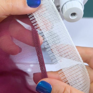 May include: A close-up of a white fabric tape with a grid pattern and small teeth along one edge. The tape is being sewn to a burgundy fabric with a sewing machine.