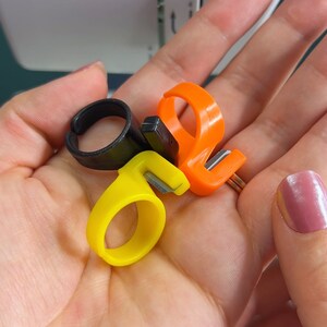 May include: Three finger thread cutters in black, orange, and yellow. The cutters have a small blade for trimming thread.