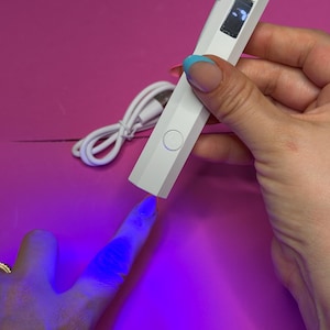May include: A white, pen-shaped nail lamp with a small screen and a USB cable. The lamp is being used to cure nail polish on a finger, illuminating it with a blue light. The background is a bright pink surface.