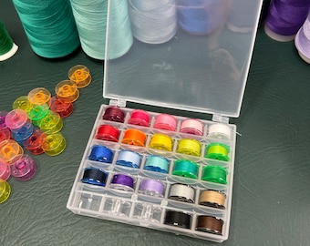 May include: A clear plastic storage box with 24 small spools of thread in various colors. The spools are arranged in rows and columns. The box is sitting on a green surface.