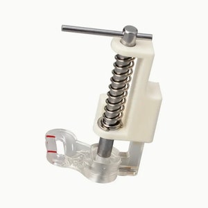 May include: A white plastic sewing machine foot with a clear plastic base and a spring-loaded metal arm. The foot is designed to be used for sewing on thick fabrics.