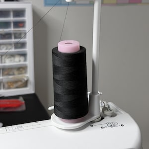 May include: A white plastic thread stand attached to a sewing machine, featuring a vertical arm that guides a single black thread upward from a large spool. The spool is mounted on a pink cylindrical core, and the stand is secured to the machine.