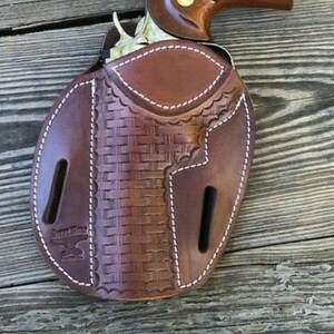 May include: A brown leather holster with a woven pattern and white stitching. A brown and silver handgun is in the holster.