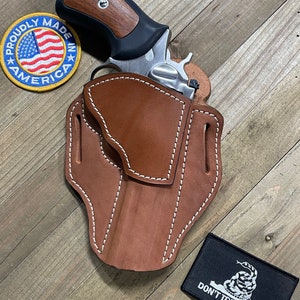 May include: A brown leather holster with white stitching, designed to hold a silver revolver with a wooden handle. The holster is on a wooden surface with a red, white, and blue "Proudly Made in America" patch and a black patch with a snake and the text "Don't Tread on Me".