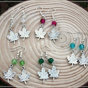 Puede incluir: Seis pares de pendientes de hoja de arce plateados con cuentas de vidrio de diferentes colores. Las cuentas son transparentes, rosadas, verdes y azules.