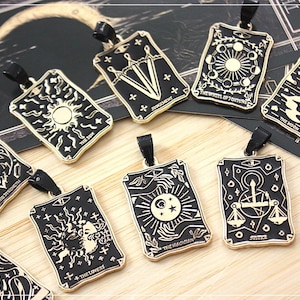 Può includere: Una collezione di pendenti neri e dorati a forma di carte dei tarocchi con vari disegni, tra cui il sole, la luna e bilance. Ogni pendente ha un nastro nero per essere indossato. I pendenti sono esposti su una superficie di legno.