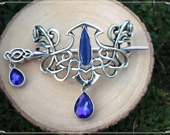 May include: A silver hair slide featuring Celtic knot designs and a central, oval-shaped blue gemstone. Two blue teardrop gems hang from the slide. The slide is presented on a light brown wooden surface.