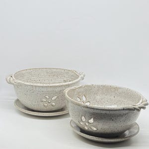 May include: Two speckled off-white ceramic berry bowls with matching saucers. Each bowl features a flower-shaped cutout on the side and twisted rope-style handles on the rim. The glaze has a light gray speckled texture throughout the surface.