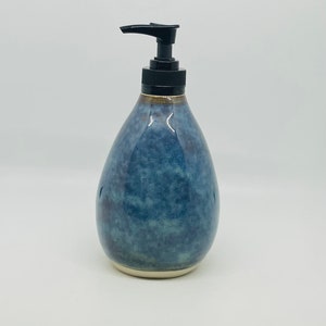 May include: A blue ceramic soap dispenser with a black pump. The dispenser has a teardrop shape and a glossy finish. The base is a light cream colour. The dispenser is designed for liquid soap or lotion.