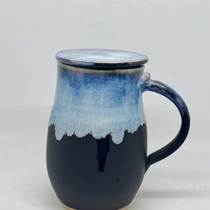 Pode incluir: Uma caneca de cerâmica com tampa, apresentando um gradiente de cores. A parte superior é azul claro, passando para branco e depois para um azul marinho escuro na base. A caneca tem uma alça curva e uma tampa correspondente.