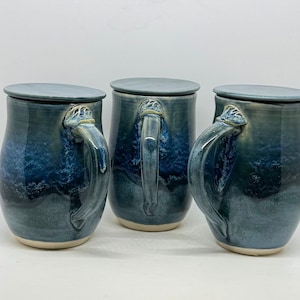 May include: Three ceramic mugs with matching lids, each featuring a rounded body and a curved handle. The glaze displays a gradient of deep navy blue to lighter slate blue, with a textured, mottled finish. The base of each mug is unglazed.