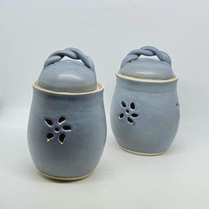 Może przedstawiać: Dwa pasujące niebieskie ceramiczne słoiki z pokrywkami. Każdy słoik ma wycięty wzór w kształcie kwiatu i skręcony uchwyt na pokrywce. Słoiki są w kolorze jasnoniebieskim z lekko teksturowaną powierzchnią i kremowym obramowaniem.