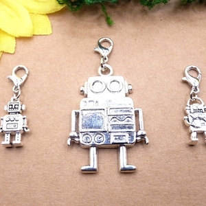 Könnte beinhalten: Drei silberfarbene Roboter-Anhänger mit Karabinerverschlüssen. Der zentrale Roboter ist größer, mit detailliertem Körper und Armen. Die anderen beiden sind kleiner, mit vereinfachten Roboterdesigns. Diese Anhänger eignen sich für die Schmuckherstellung oder zum Basteln.