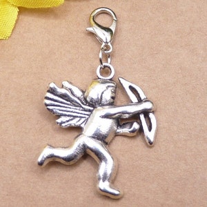 Könnte beinhalten: Ein silberfarbener Anhänger, der Amor, den römischen Liebesgott, mit Flügeln und Pfeil und Bogen darstellt. Der Anhänger ist an einem silberfarbenen Verschluss befestigt, der für ein Armband oder eine Halskette geeignet ist. Der Anhänger ist aus Metall und hat ein detailliertes Design.
