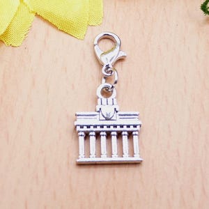 K&ouml;nnte beinhalten: Silberfarbener Charm-Anh&auml;nger, der ein Geb&auml;ude mit S&auml;ulen darstellt, wahrscheinlich ein Wahrzeichen. Der Anh&auml;nger ist an einem Verschluss befestigt. Der Hintergrund ist eine helle Holzoberfl&auml;che.