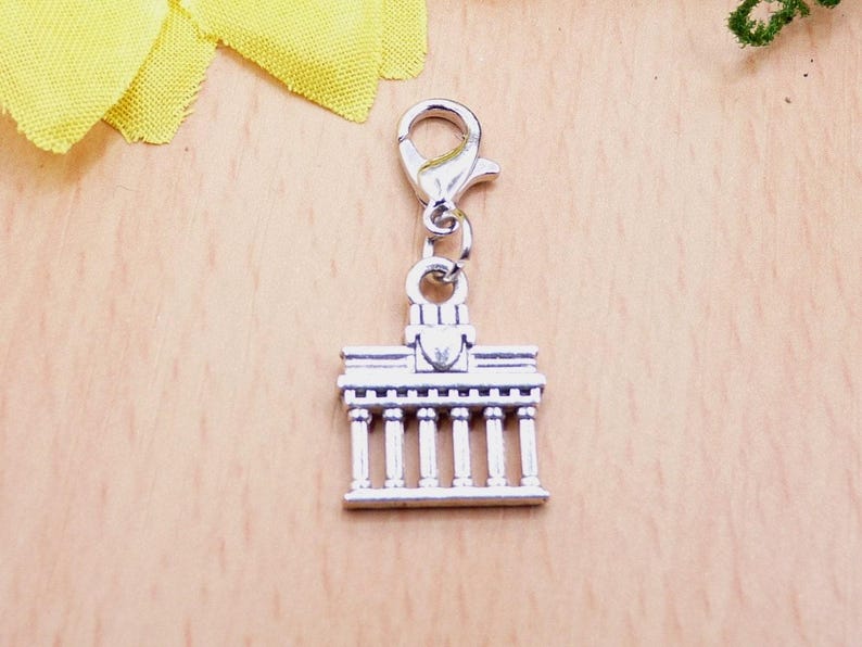K&ouml;nnte beinhalten: Silberfarbener Charm-Anh&auml;nger, der ein Geb&auml;ude mit S&auml;ulen darstellt, wahrscheinlich ein Wahrzeichen. Der Anh&auml;nger ist an einem Verschluss befestigt. Der Hintergrund ist eine helle Holzoberfl&auml;che.