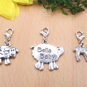 Könnte beinhalten: Drei silberfarbene Schaf-Anhänger mit Karabinerverschlüssen. Ein Anhänger trägt den Text "Bella Baby". Die Anhänger haben die Form eines Schafs, eines Lamms und eines Schafs mit dem Text "Bella Baby".