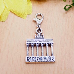 K&ouml;nnte beinhalten: Silberfarbener Charm-Anh&auml;nger mit dem Brandenburger Tor und dem Wort "BERLIN" am unteren Rand. Der Anh&auml;nger hat einen Karabinerverschluss. Der Hintergrund ist eine helle Holzoberfl&auml;che mit einer gelben Blume und gr&uuml;nen Bl&auml;ttern.