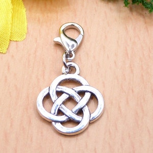 Könnte beinhalten: Ein silberfarbener Anhänger mit einem keltischen Knotenmuster, befestigt an einem Karabinerverschluss. Der Anhänger weist ein detailliertes, ineinander verwobenes Muster auf und besteht aus einem glänzenden, metallischen Material. Der Hintergrund ist eine hellbraune Holzoberfläche.