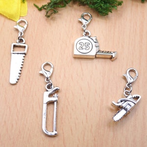 Könnte beinhalten: Silberfarbene Charms mit Karabinerverschlüssen, die Miniaturwerkzeuge darstellen. Die Sammlung umfasst eine Säge, ein Maßband mit der Zahl 25, einen Schraubenschlüssel und eine Kettensäge. Diese Charms sind ideal für Schmuckherstellung oder Bastelprojekte.