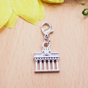 K&ouml;nnte beinhalten: Silberfarbener Charm mit einer Miniatur eines Geb&auml;udes mit S&auml;ulen, befestigt an einem silberfarbenen Karabinerverschluss. Der Charm ist wahrscheinlich ein Souvenir oder ein Schmuckbestandteil. Der Hintergrund ist eine helle Holzoberfl&auml;che mit einer gelben Blume.