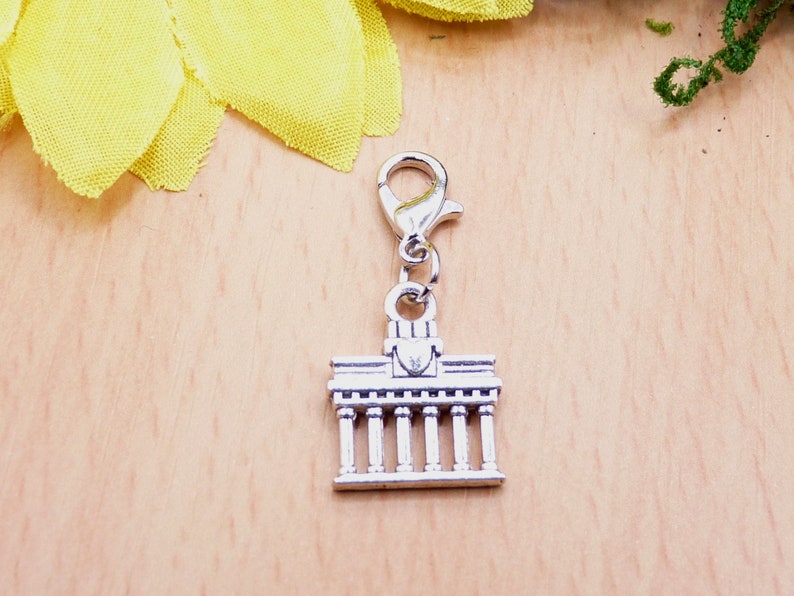 K&ouml;nnte beinhalten: Silberfarbener Charm mit einer Miniatur eines Geb&auml;udes mit S&auml;ulen, befestigt an einem silberfarbenen Karabinerverschluss. Der Charm ist wahrscheinlich ein Souvenir oder ein Schmuckbestandteil. Der Hintergrund ist eine helle Holzoberfl&auml;che mit einer gelben Blume.