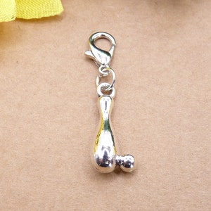 Könnte beinhalten: Ein silberfarbener Charm-Anhänger in Form einer Bowlingkegel mit einer kleinen Kugel. Der Anhänger hat einen Karabinerverschluss zur einfachen Befestigung an einem Armband oder einer Halskette. Der Anhänger ist aus glänzendem, reflektierendem Metall gefertigt.