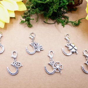 Könnte beinhalten: Silberfarbene Anhänger mit Karabinerverschlüssen, die Hufeisen- und vierblättriges Kleeblatt-Designs aufweisen. Die Anhänger werden auf einer hellbraunen Oberfläche mit gelben Sonnenblumen und grünem Moos im Hintergrund präsentiert.