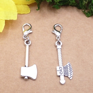 Könnte beinhalten: Zwei silberfarbene Axt-Anhänger mit Karabinerverschluss. Ein Axt-Anhänger hat eine einfache Klinge, der andere ein detailliertes Griff- und Klingendesign. Geeignet für die Schmuckherstellung oder Bastelarbeiten.