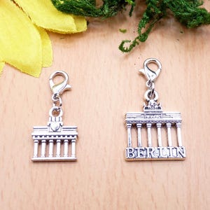 K&ouml;nnte beinhalten: Zwei silberfarbene Berlin Brandenburger Tor Charms mit Karabinerhaken. Ein Charm hat das Wort "BERLIN" eingraviert. Die Charms sind detailliert mit S&auml;ulen und architektonischen Merkmalen, ideal f&uuml;r Schmuckherstellung oder als Souvenir.