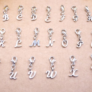Könnte beinhalten: Eine Sammlung silberfarbener Alphabet-Anhänger, jeweils mit einem Karabinerverschluss. Die Buchstaben A bis Z sind abgebildet und zeigen eine Vielzahl von Schreib- und Blockschriftarten. Die Anhänger sind auf einer hellbraunen Oberfläche angeordnet.