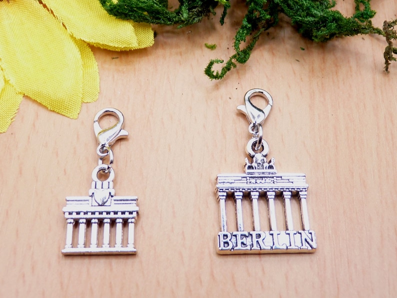 K&ouml;nnte beinhalten: Zwei silberfarbene Anh&auml;nger mit dem Brandenburger Tor. Einer ist eine kleinere, vereinfachte Version, der andere tr&auml;gt den Schriftzug "BERLIN" darunter. Beide Anh&auml;nger haben einen Karabinerverschluss.