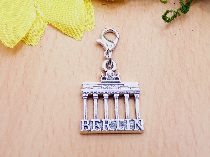 K&ouml;nnte beinhalten: Silberfarbener Charm-Anh&auml;nger mit dem Brandenburger Tor und dem Wort "BERLIN" am Sockel. Der Anh&auml;nger hat einen Karabinerverschluss zur Befestigung. Das Design ist detailliert, und der Anh&auml;nger ist wahrscheinlich aus Metall.