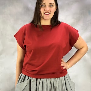 Könnte beinhalten: Eine Frau trägt ein rotes Oberteil mit kurzen Ärmeln und einen grauen Rock mit Taschen.