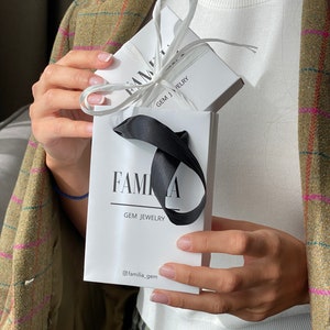 May include: A white gift bag with a black ribbon and the text "Familia Gem Jewelry" and the social media handle "@familia_gem". The bag is holding a white box with a black ribbon and the text "Familia Gem Jewelry".