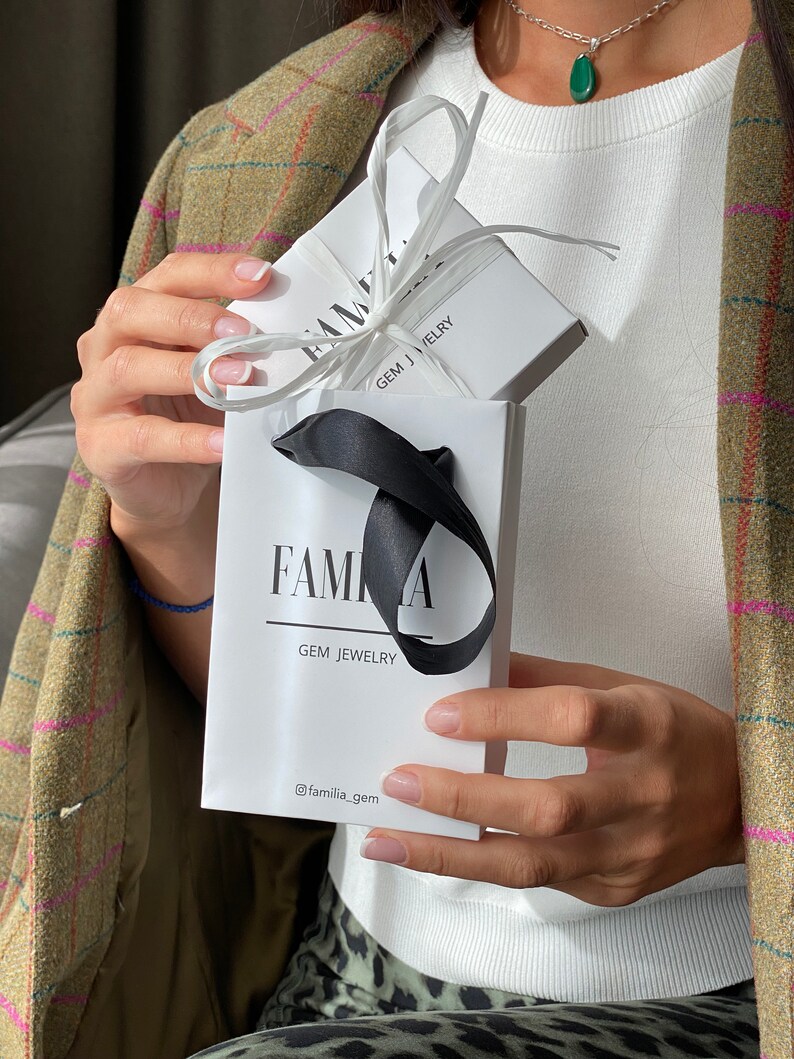 May include: A white gift bag with a black ribbon and the text "Familia Gem Jewelry" and the social media handle "@familia_gem". The bag is holding a white box with a black ribbon and the text "Familia Gem Jewelry".