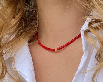 May include: A red coral necklace with a gold bead accent. The necklace is worn by a person with long blonde hair.