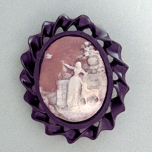 May include: A vintage cameo brooch depicting a woman and a deer in a landscape. The oval cameo is set in a dark, decorative frame. The woman is wearing a long dress and has her arm outstretched. A classic piece of jewellery.
